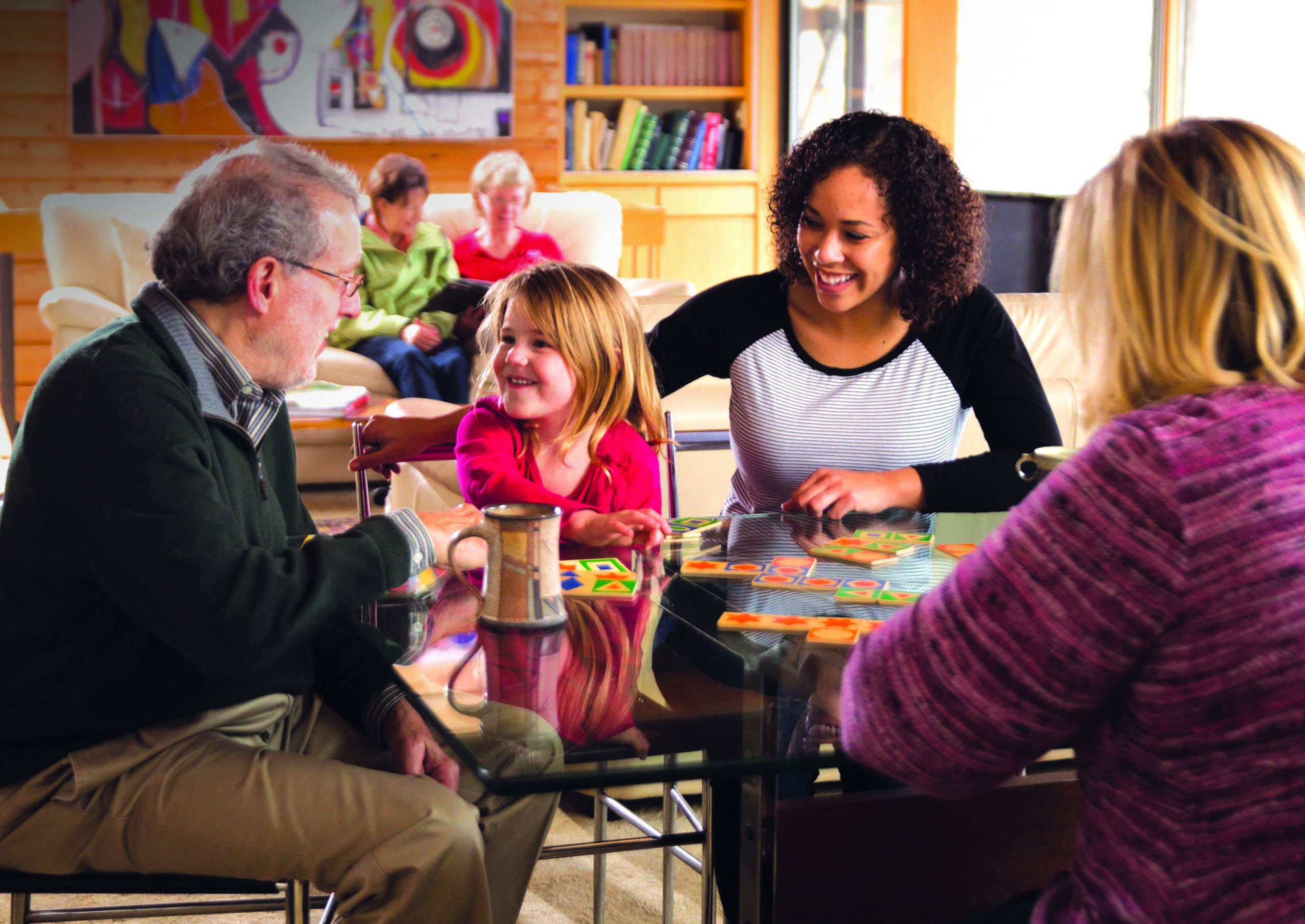 Four people around a table - an older man, young girl, mid-20s female and older woman - all are looking at the smiling child who is playing with blocks.